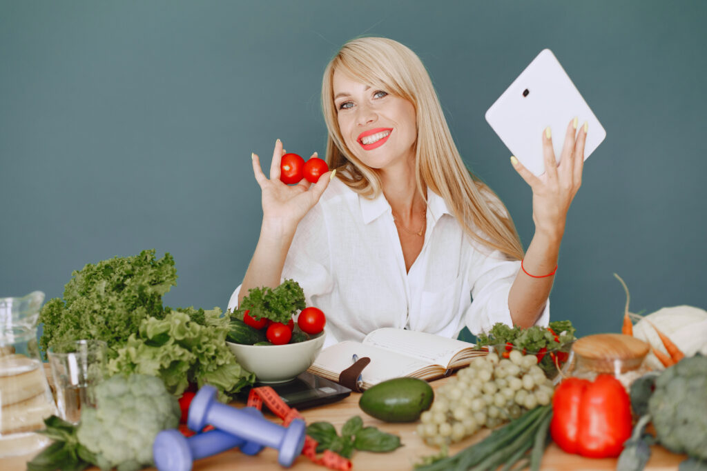 Mulher sorridente segurando tomates e uma tabela de alimentos termogênicos com vegetais ao redor.