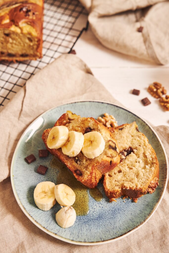 Fatias de bolo de banana com aveia servidas em um prato azul, decoradas com rodelas de banana e mel, com pedaços de chocolate e nozes ao redor.