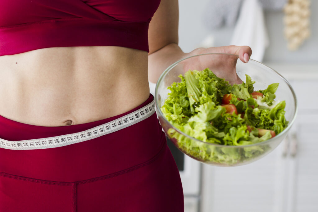 Mulher segurando salada com fita métrica na cintura, simbolizando perda de peso com alimentação saudável.