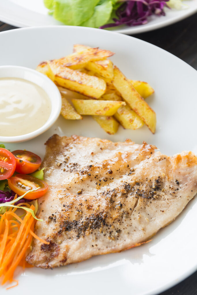 Filé de peixe assado com batata-doce, acompanhado de salada de tomate cereja e cenoura ralada, servido com molho branco em um prato branco.