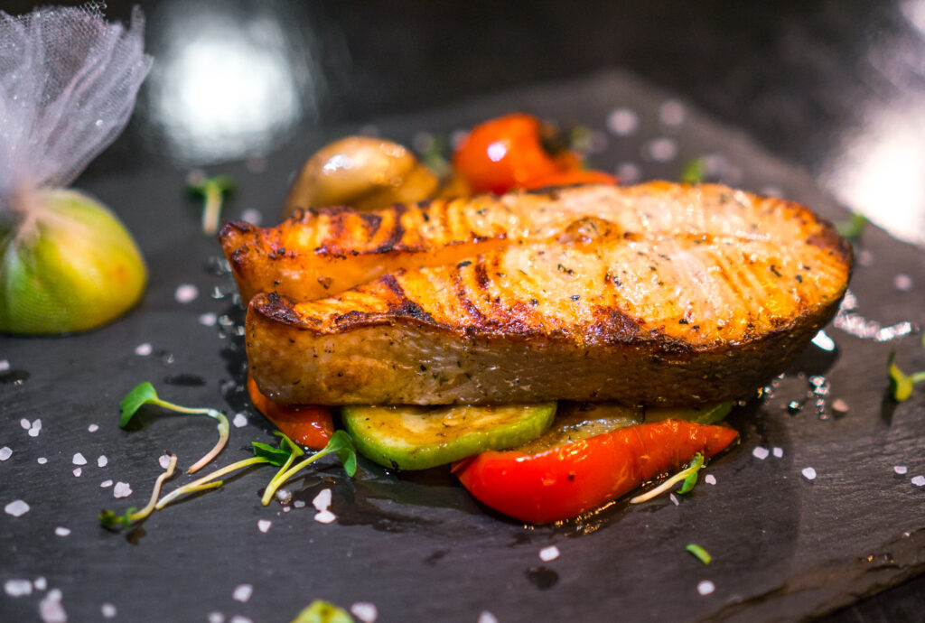 Filé de peixe grelhado servido sobre legumes assados, acompanhado de um limão decorado e finalizado com brotos e sal grosso.