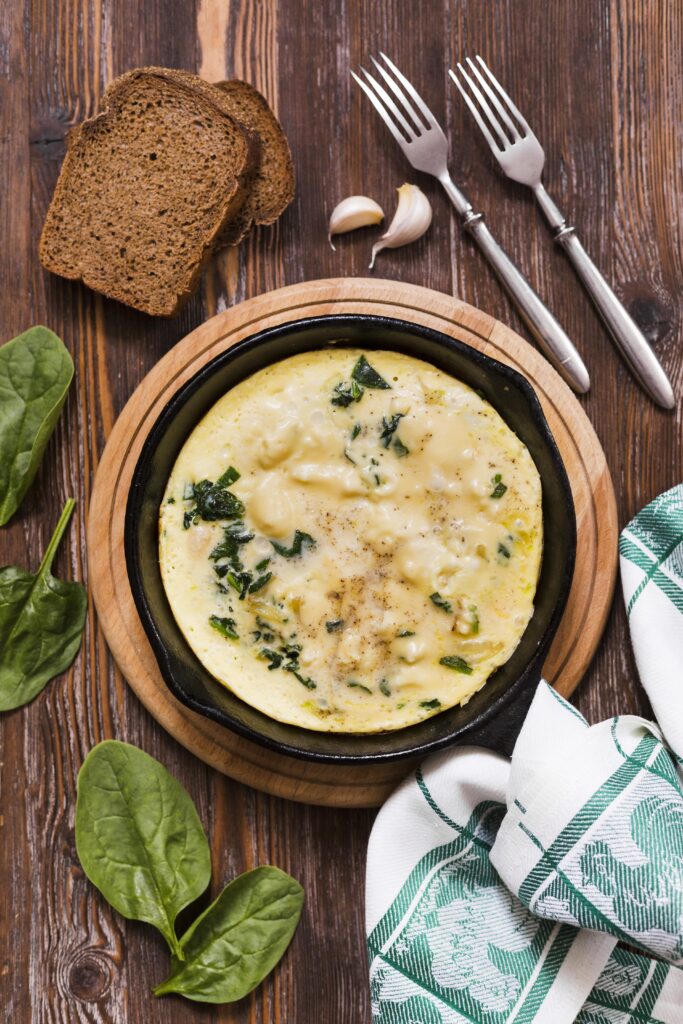 Omelete de espinafre com queijo cottage servido em uma frigideira preta, acompanhado de fatias de pão integral e talheres sobre uma mesa de madeira.