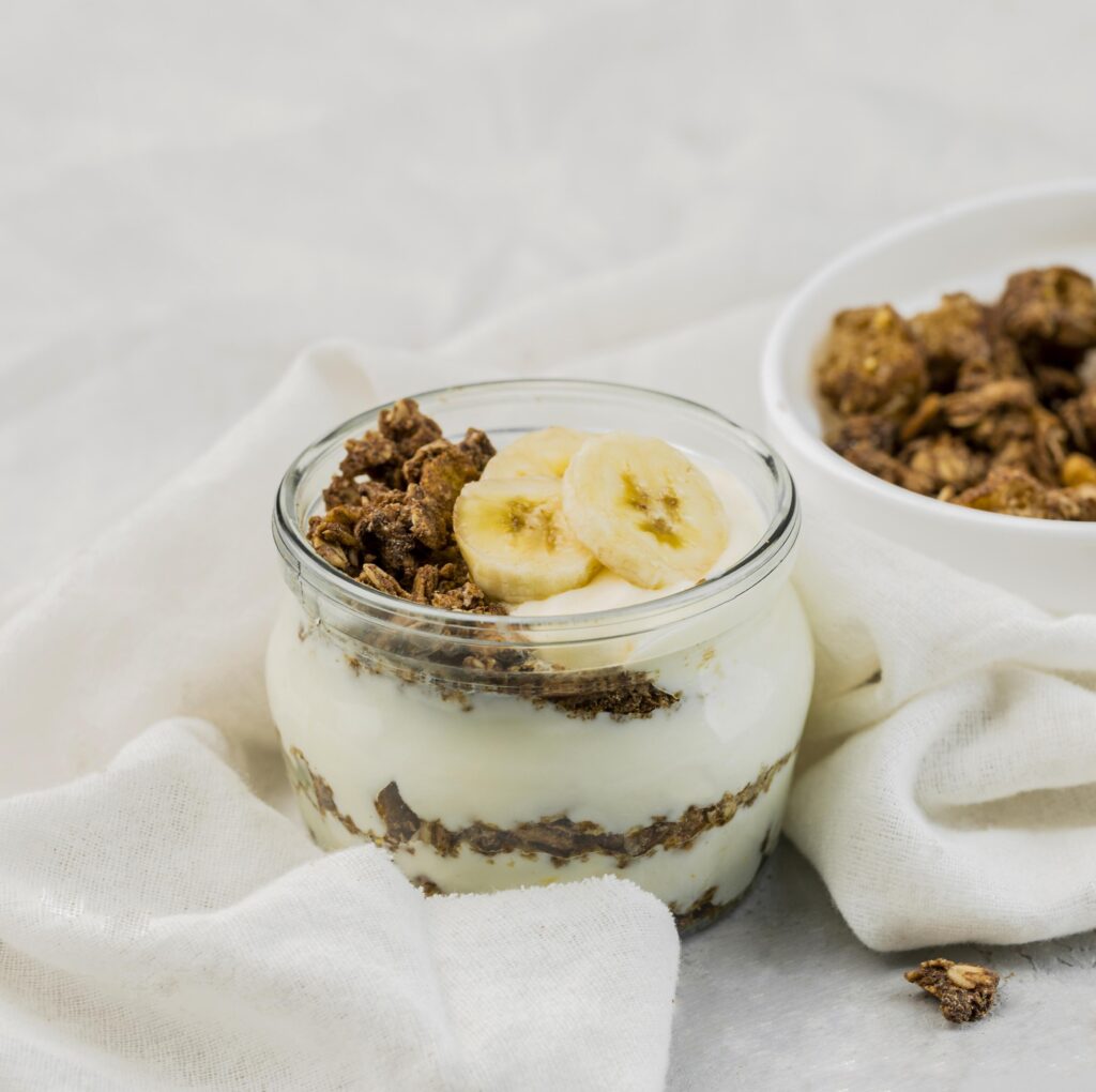 Pudim de chia com leite de coco em um pote de vidro, decorado com rodelas de banana e granola crocante, sobre um fundo branco claro.