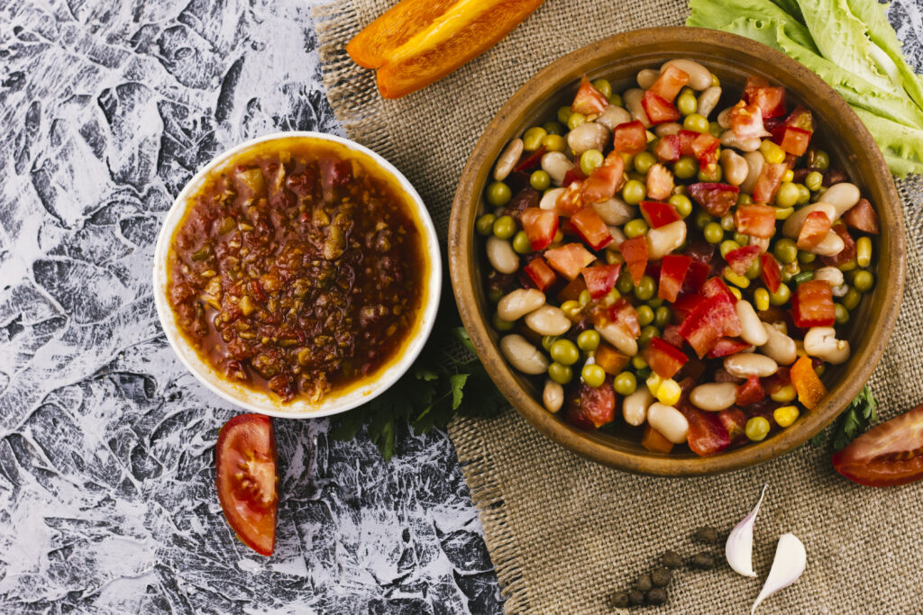 Tigela de quinoa com legumes salteados e uma tigela de molho vermelho espesso, acompanhadas de pedaços de tomate, pimentão e alho sobre uma superfície rústica.