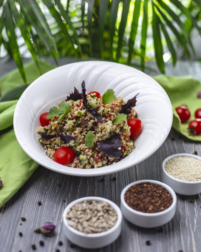 Salada de quinoa com tomates cereja, pepino, manjericão roxo e folhas de salsa servida em um prato branco, com sementes variadas ao lado, sobre uma mesa de madeira, com uma planta verde ao fundo.