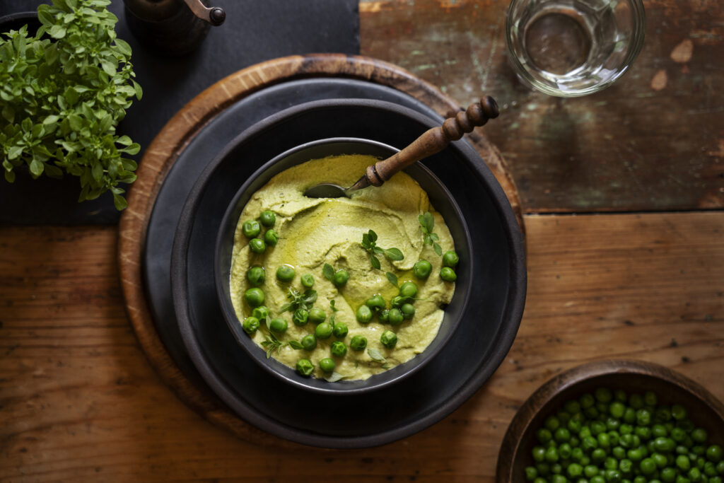 Sopa fria de pepino e hortelã servida em uma tigela preta, decorada com ervilhas frescas e ervas, com colher de madeira ao lado, sobre uma mesa rústica de madeira.