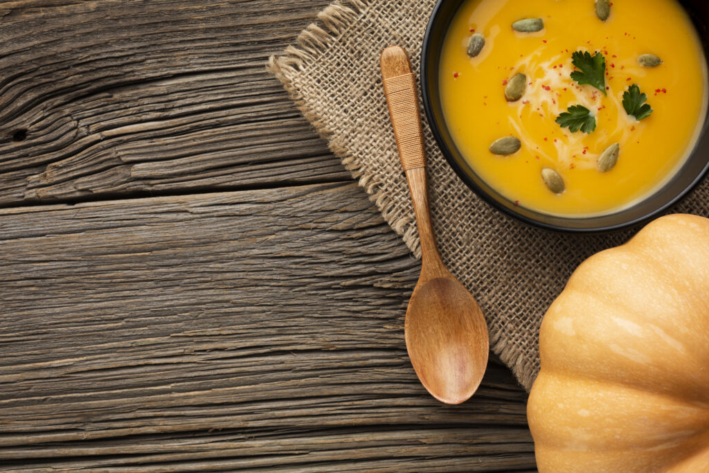 Tigela de sopa de abóbora com gengibre, decorada com sementes de abóbora e ervas frescas, servida sobre um pano rústico com uma colher de madeira ao lado.