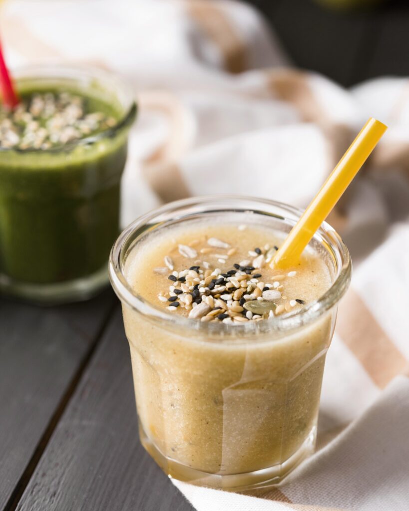 Smoothie de banana com canela servido em um copo de vidro com canudo amarelo, decorado com sementes, acompanhado de um smoothie verde ao fundo.