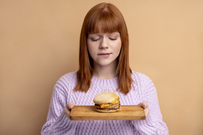 Mulher segurando um hambúrguer com expressão contemplativa, focada em controlar a fome emocional.