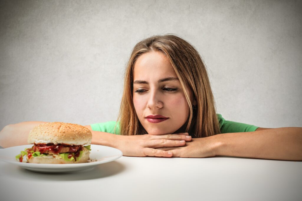 Mulher olhando para um hambúrguer com expressão de reflexão sobre fome emocional.