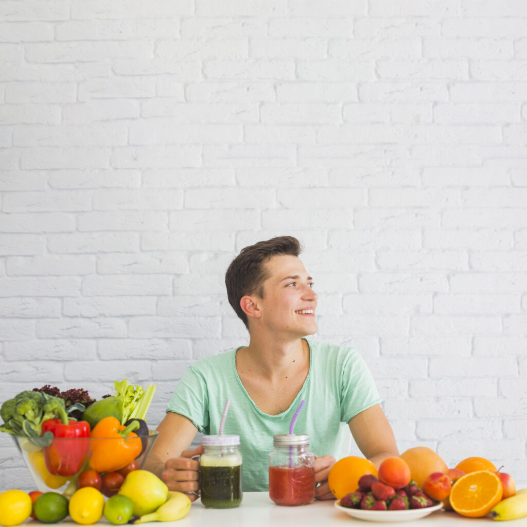 Homem sorrindo com mesa repleta de frutas e vegetais, seguindo uma dieta vegetariana.