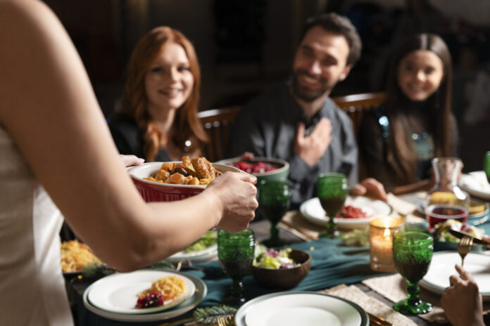 Pessoa servindo um prato de comida durante um jantar, com convidados sorrindo ao redor da mesa decorada com taças verdes e velas.