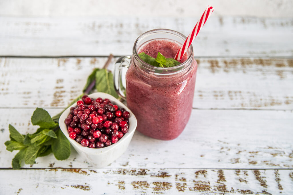 Smoothie de morango servido em um copo de vidro com canudo listrado, acompanhado de uma tigela de frutas vermelhas congeladas e folhas de hortelã.