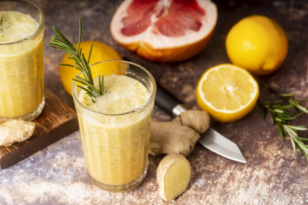 Suco detox de gengibre servido em copo de vidro com raminho de alecrim, acompanhado de limões, toranja e gengibre fresco sobre a mesa.