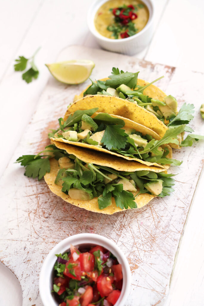 Tacos de alface com frango grelhado, recheados com folhas verdes e abacate, servidos em uma tábua de madeira, acompanhados de molho e salsa fresca.