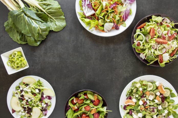 Variedade de saladas refeição ricas em proteínas dispostas em pratos, com ingredientes frescos como folhas, legumes e frutas, em uma mesa escura.