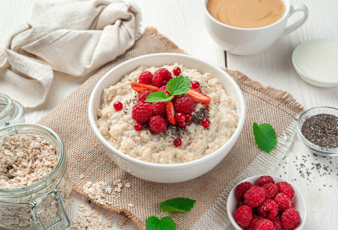 Tigela de mingau de aveia com frutas vermelhas frescas, chia e hortelã, acompanhada por itens como aveia, sementes e café.