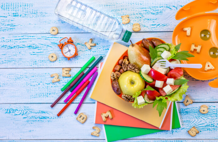 Lancheira escolar saudável com frutas, vegetais, queijos e nozes, ao lado de materiais escolares coloridos e uma garrafa de água.