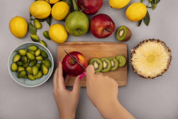 Pessoa cortando maçã com kiwis, limões, maçãs e abacaxi ao redor em uma mesa, destacando frutas ideais para perder peso.