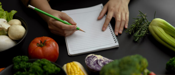 Pessoa escrevendo em um caderno cercado por vegetais frescos, incluindo tomate, brócolis e cogumelos, simbolizando o planejamento de refeições veganas saudáveis.