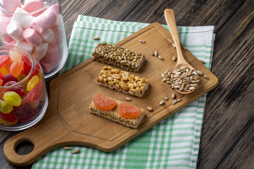 Barras proteicas caseiras com diferentes ingredientes, servidas em uma tábua de madeira ao lado de sementes e guloseimas.