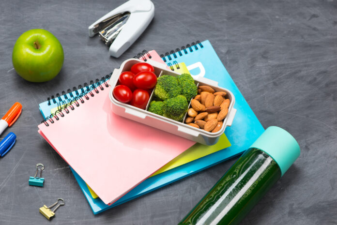 Lancheira saudável com tomate-cereja, brócolis e amêndoas sobre cadernos coloridos em uma mesa com itens de escritório.