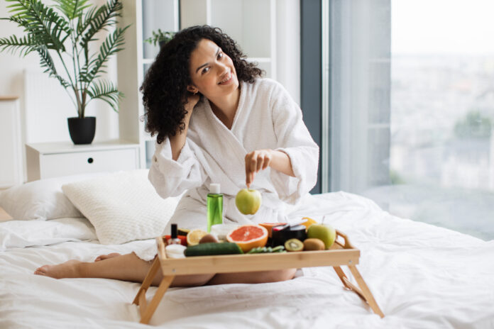 Mulher sorridente comendo frutas saudáveis na cama enquanto desfruta de sua rotina matinal de bem-estar.