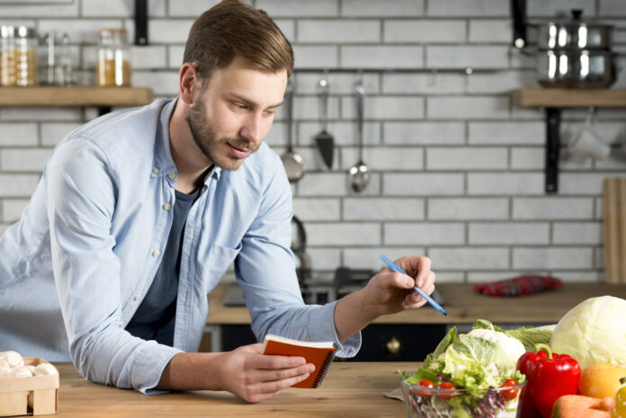 Homem em uma cozinha planejando refeições com caderno e lápis, cercado por vegetais frescos.