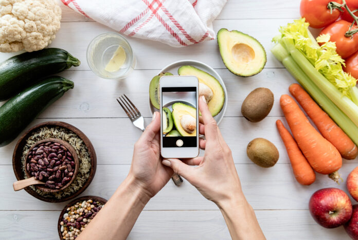 Mão segurando um celular fotografando um prato de abacate, rodeado por legumes frescos como cenouras, tomates e abobrinha, representando planejamento de refeições low carb.