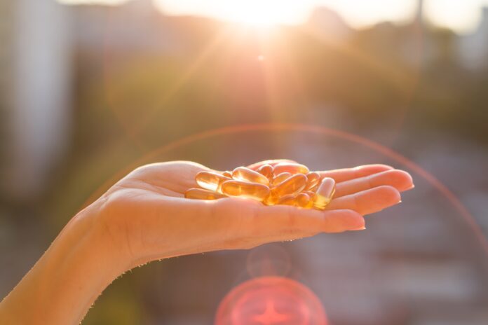 Mão segurando cápsulas de vitamina D iluminadas pelo sol, destacando a relação entre suplementação e bem-estar.