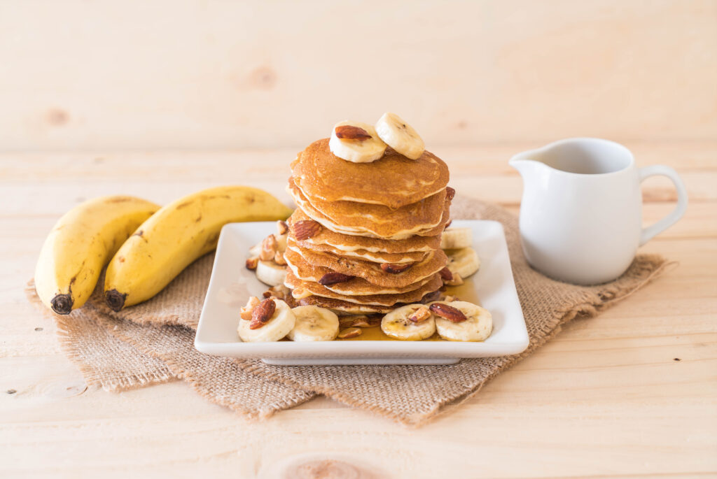 Panquecas de aveia com banana decoradas com fatias de banana, amêndoas e mel, servidas em um prato branco, acompanhadas de bananas frescas e uma jarra de leite.