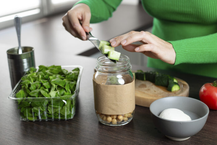 Mulher preparando uma salada em pote com grão-de-bico, pepino e folhas verdes em uma cozinha.