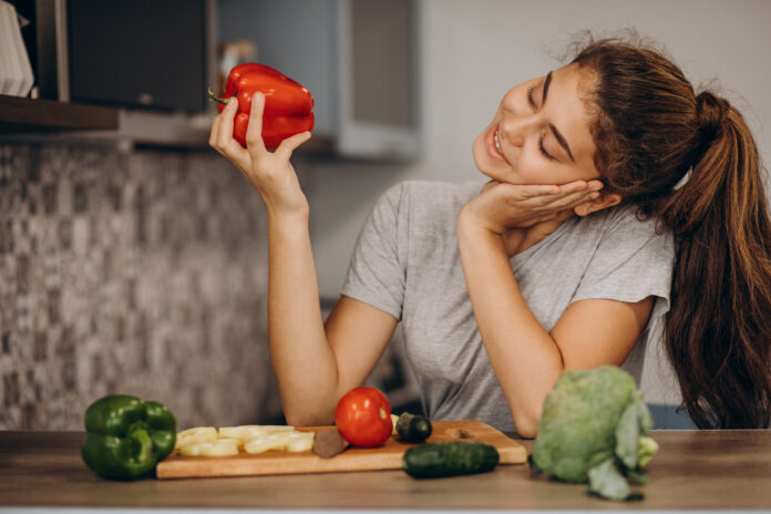 Mulher sorrindo enquanto segura um pimentão vermelho, rodeada de vegetais frescos em uma cozinha.