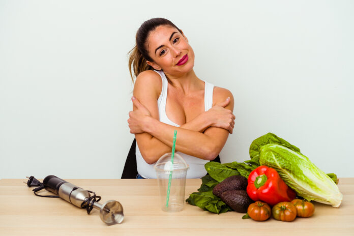 Mulher feliz abraçando a si mesma ao lado de vegetais frescos e um copo de smoothie, representando uma dieta flexível e saudável.