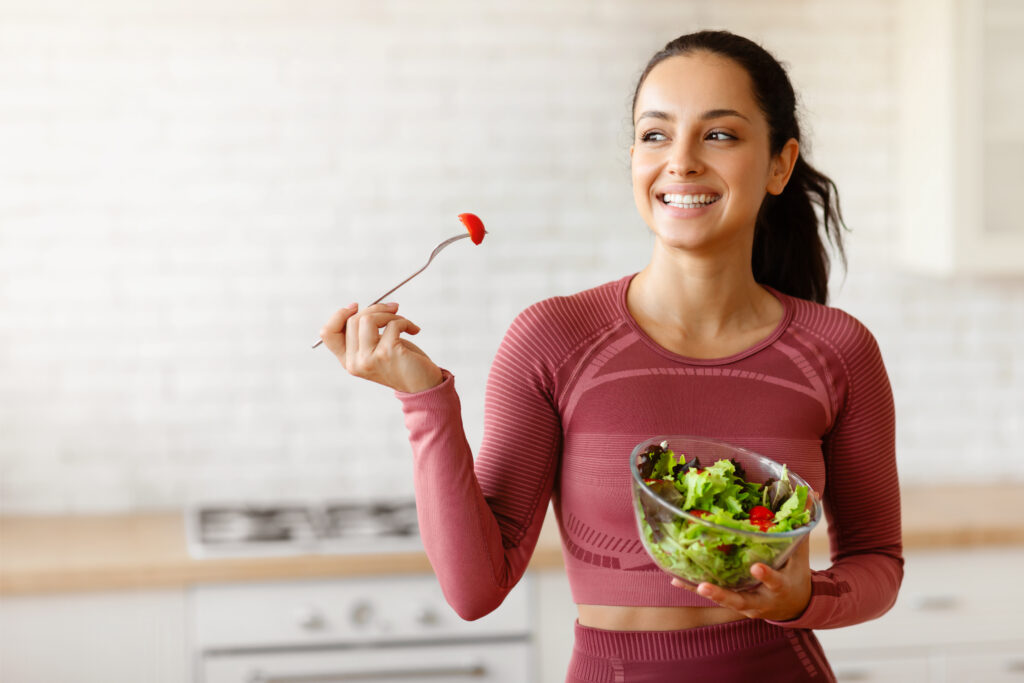 Mulher sorrindo enquanto segura uma tigela de salada e um garfo com tomate, representando alimentação saudável e dieta flexível.