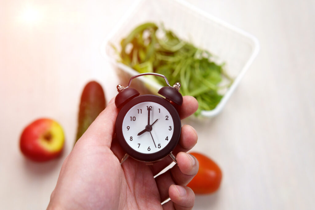 Mão segurando um relógio, com alimentos saudáveis ao fundo, representando o conceito de dieta intermitente.