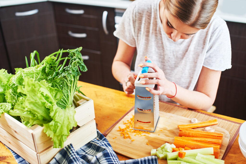 Mulher ralando cenoura em uma cozinha organizada com vegetais frescos ao lado.