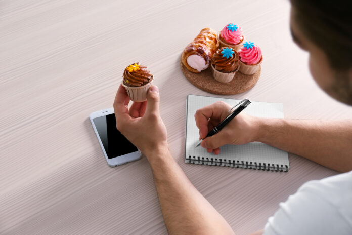 Pessoa escrevendo um plano alimentar enquanto segura um cupcake, com outros doces e um celular sobre a mesa.