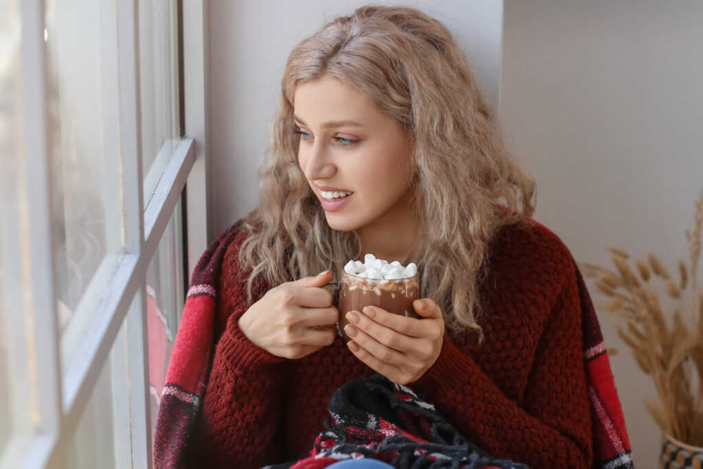 Mulher segurando uma xícara de chocolate quente com marshmallows, olhando pela janela enquanto reflete sobre hábitos alimentares saudáveis.