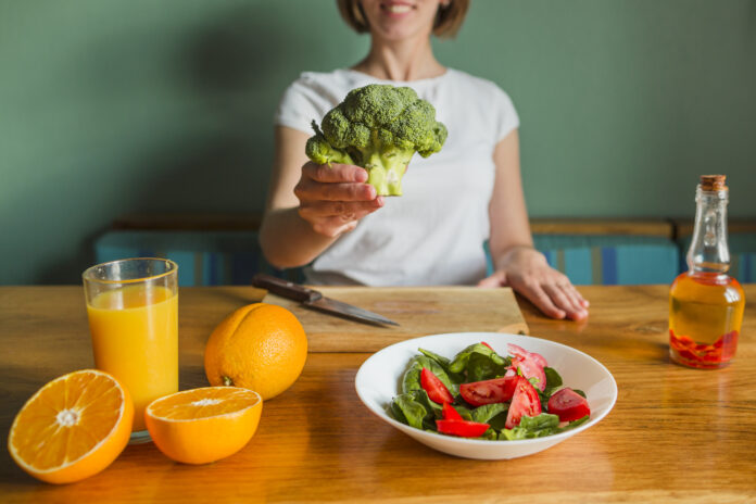 Pessoa segurando um brócolis ao lado de suco de laranja, frutas cítricas, salada de espinafre e tomate, representando uma dieta anti-inflamatória saudável.