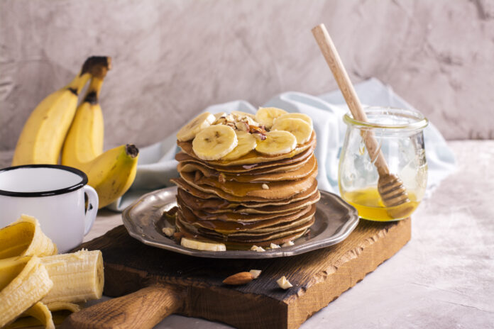 Panquecas de aveia com banana decoradas com fatias de banana e mel, ao lado de bananas frescas e um pote de mel.