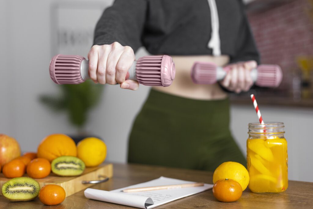 Pessoa segurando halteres com frutas, suco natural e bloco de notas ao fundo, representando a conexão entre exercícios e alimentação saudável.