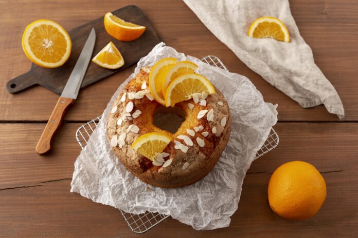 Bolo integral de laranja com chia decorado com fatias de laranja e amêndoas, servido sobre papel manteiga em uma grade de resfriamento.