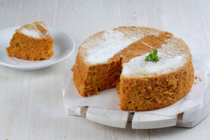 Bolo de batata doce com coco sem glúten e sem açúcar, decorado com açúcar de confeiteiro e folhas verdes, com uma fatia servida em um prato branco.