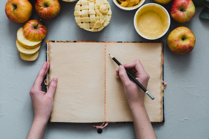 Mãos escrevendo em um diário alimentar com alimentos frescos ao redor.