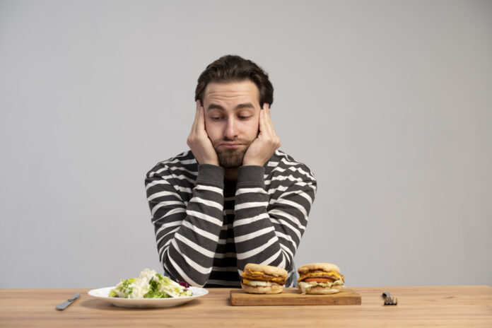 Homem pensativo avaliando entre uma refeição saudável e hambúrgueres, representando o dilema da fome emocional.