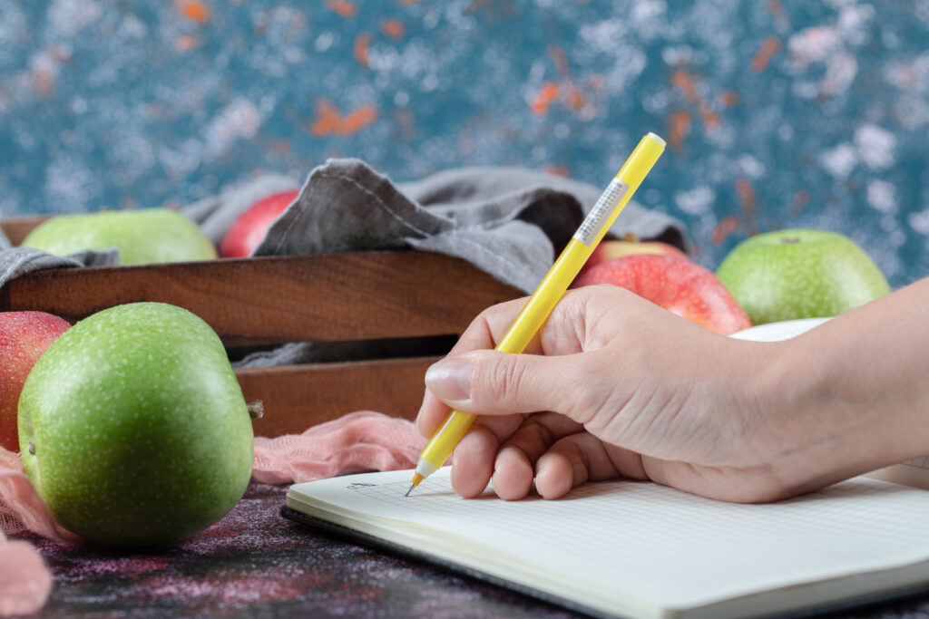 Mão segurando uma caneta amarela, escrevendo em um diário alimentar, com maçãs frescas ao fundo.