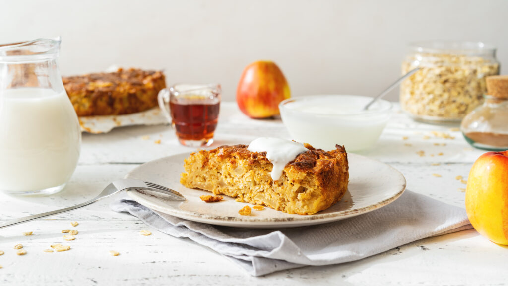 Fatia de bolo de aveia com iogurte grego e mel, acompanhada de ingredientes saudáveis em um fundo iluminado.