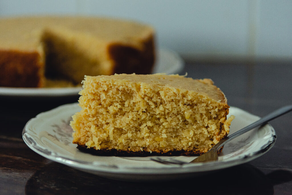 Fatia de bolo de batata doce com coco sem glúten, servida em um prato branco decorativo com o bolo inteiro ao fundo.