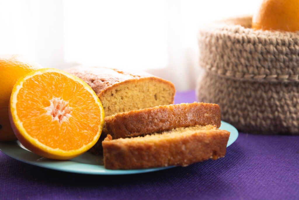 Fatias de bolo integral de laranja com chia em um prato azul, ao lado de uma laranja cortada ao meio e um cesto decorativo no fundo.